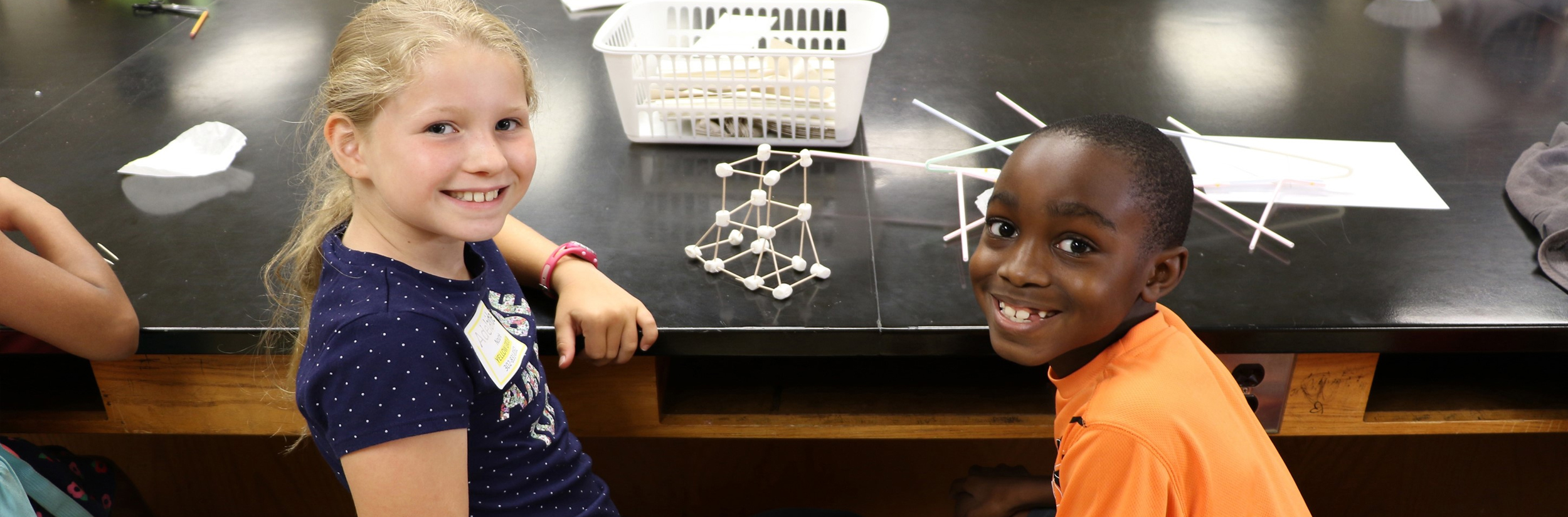 A boy and girl in elementary school smile at the camera after completing a science activity with toothpicks and marshmallows in a science classroom.