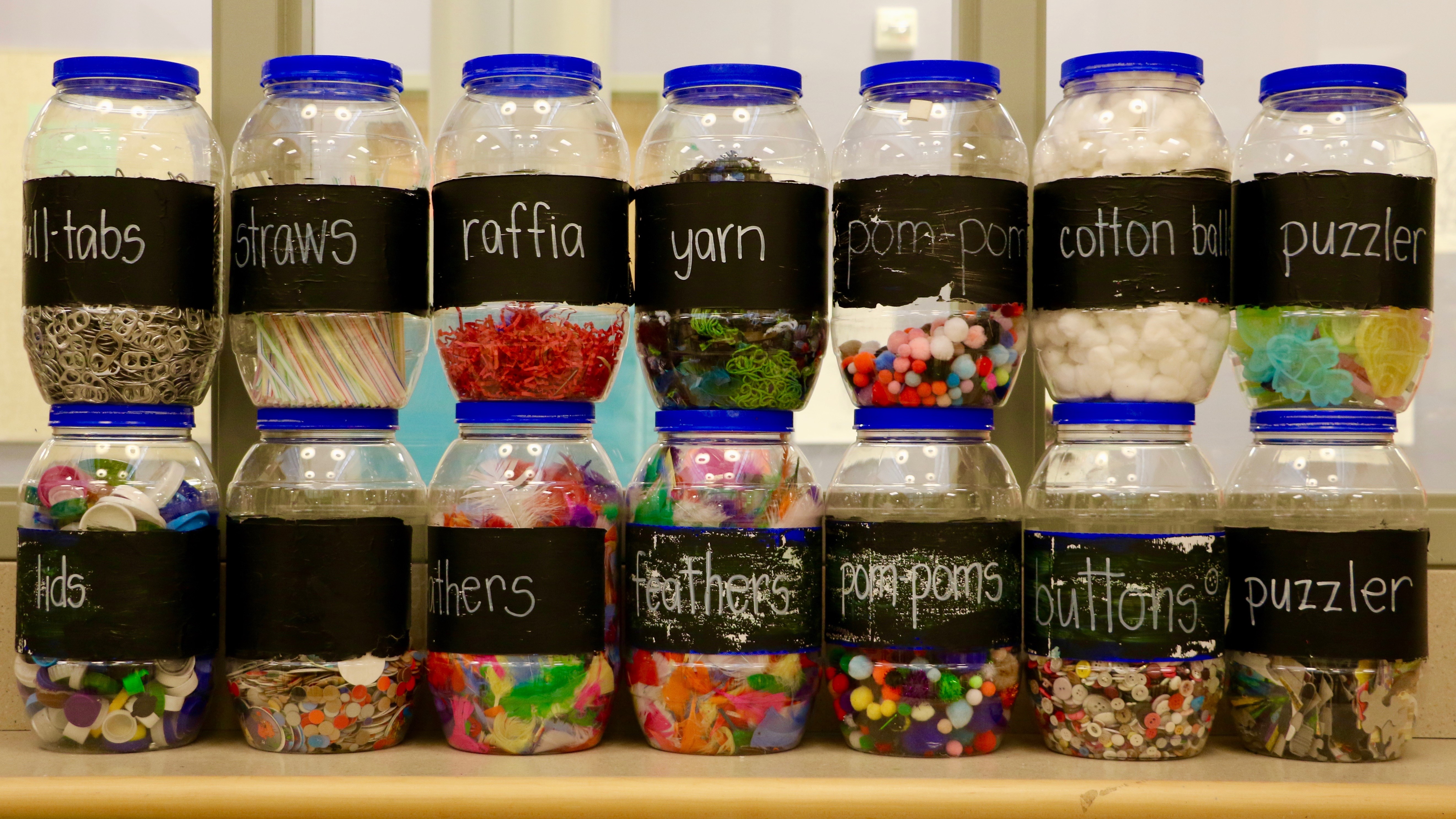 This image shows a row of labeled plastic jars with art supplies in them, including straws, buttons, cotton balls, feathers, and yarn.