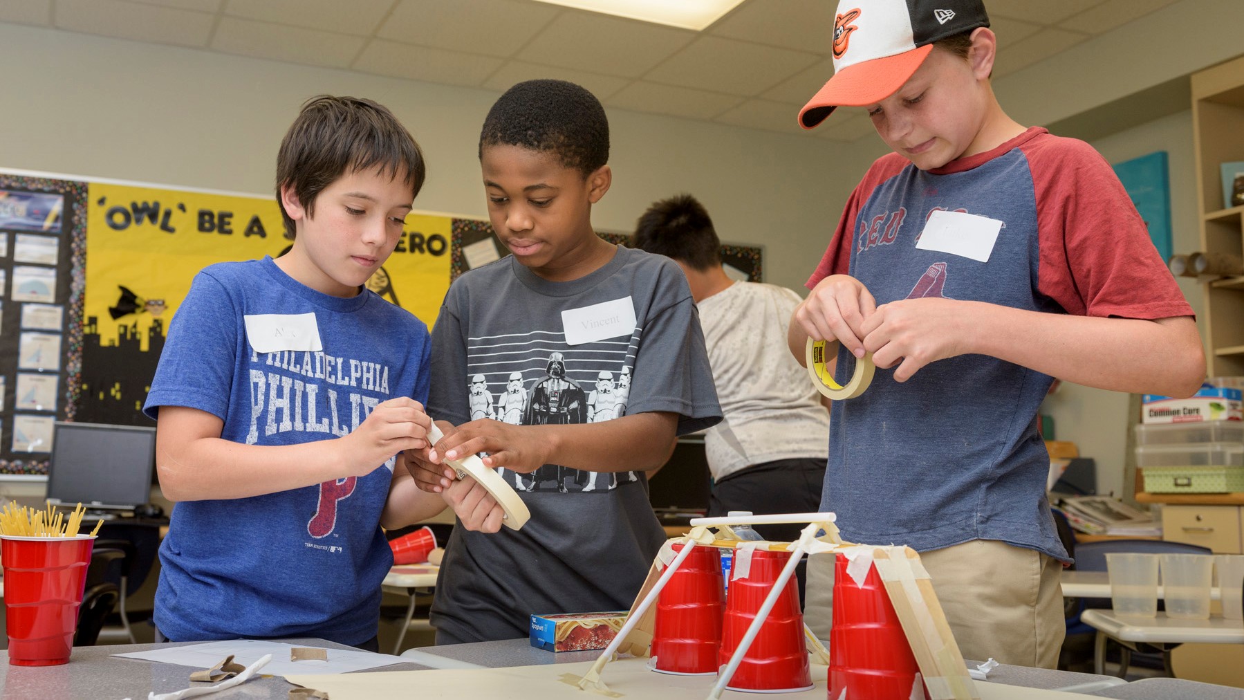 Three middle school boys engage in a STEM activity with plastic cups, spaghetti, and tape in a CEHD summer camp.
