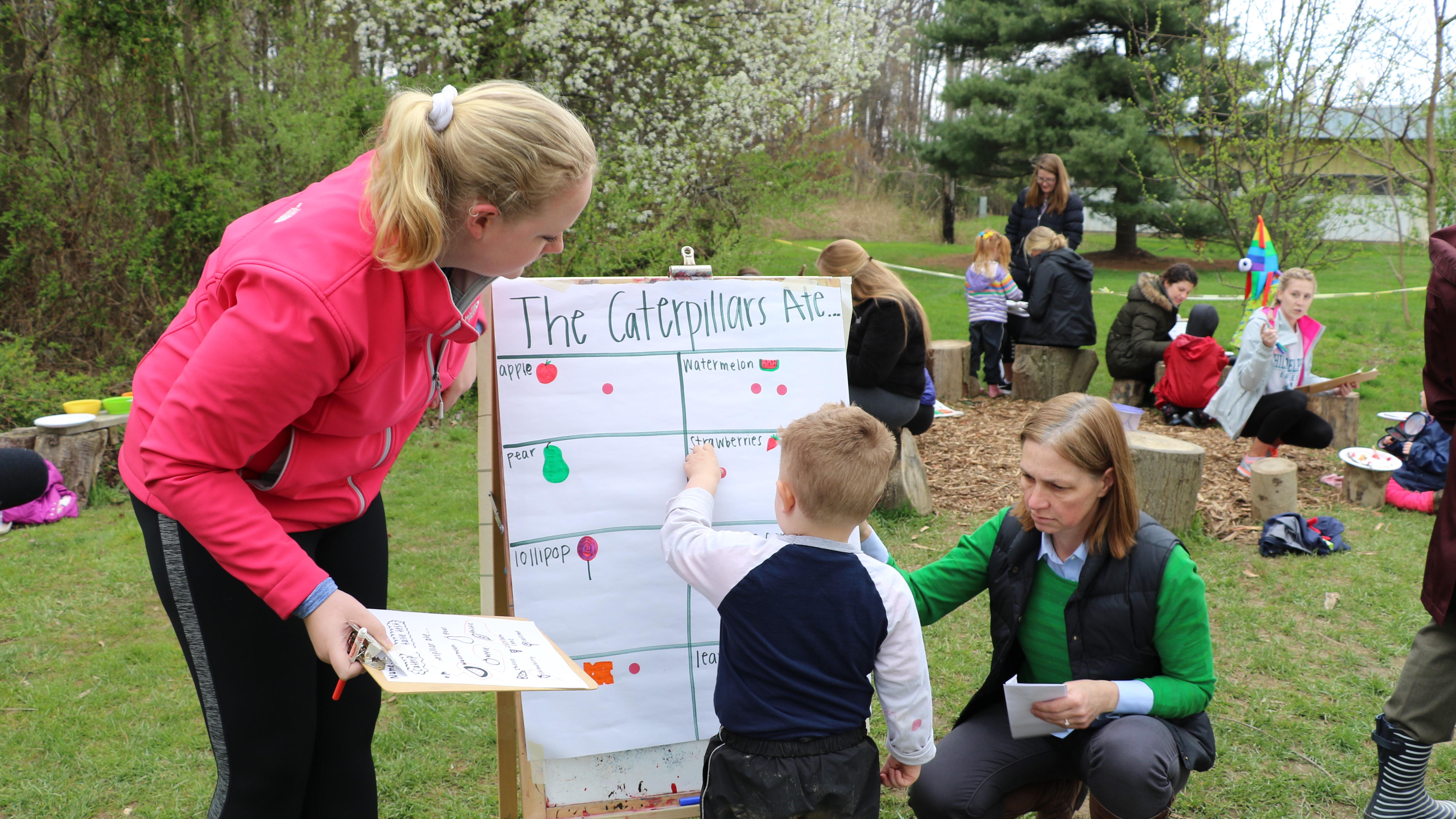 A student teacher leads a child in a science activity about caterpillars using a poster board outside.