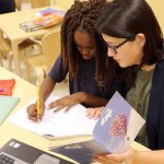 Two students work together on a literacy assignment in classroom with books, paper and pencil, and a computer.