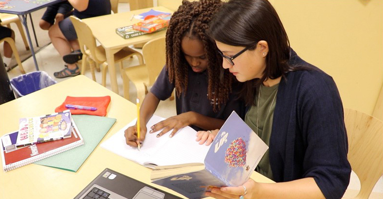 Two students work together on a literacy assignment in classroom with books, paper and pencil, and a computer.